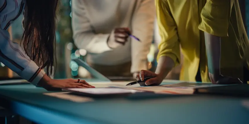 Three professionals lean over a table reviewing printed reports and documents together reflecting the collaborative campaign planning process at a results-driven marketing agency at EverConvert.